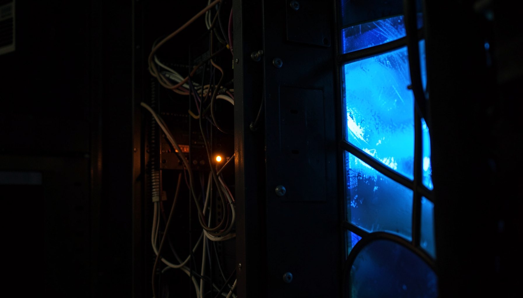 A dimly lit server rack with tangled cables and a single amber warning LED glowing against dark metal panels, moody and industrial.